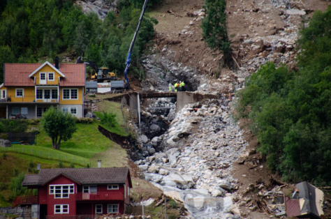 Et gult og et rødt hus ligger over hverandre i en bratt skråning, Rett til høyre for husene har det gått et stort ras og det renner en elv ned gjennom raset.